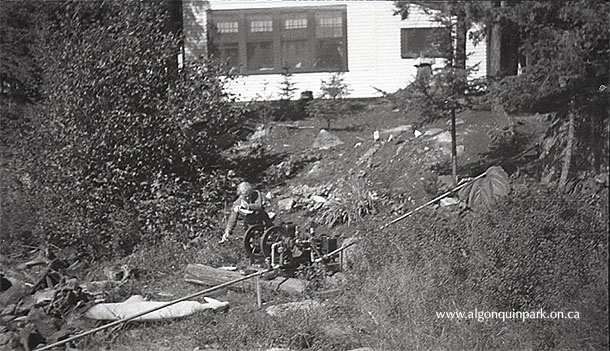 Image: A cottager at the King Leasehold on Cache Lake watches a water pump on the hillside, 1936. Pipes lead from the lake (not seen) to the cottage above. APPAC, 2002.5.183, H.M. King. 