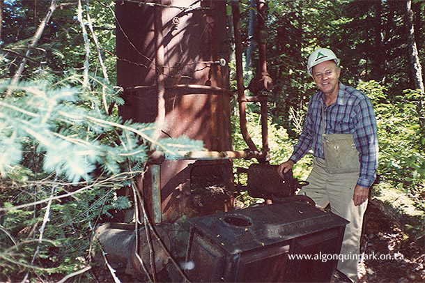 Image: Ray Townsend stands with one of the two donkey engines re-discovered near Proulx Lake, 1990. Although the wooden buildings that once sheltered the machines were long gone, other artifacts remained from the operation, including a wood stove visible in the foreground of this image. 