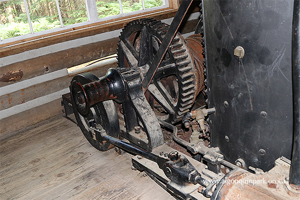 Image: Detail of the winch on the front of the donkey engine, Algonquin Logging Museum. The thick metal cable runs through an opening in the building towards the hill where it would be attached to a sleigh. 